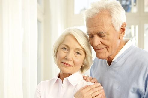 Ein älteres Paar steht nah beieinander, der Mann umarmt die Frau sanft, beide lächeln in eine ruhige Umgebung.